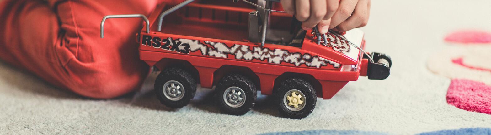 Child playing with red and black vehicle toy on light-colored carpet.