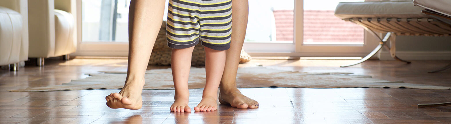 Adult standing behind child on tiled floor. The adult is seen from the thighs down. The child from the waist down.